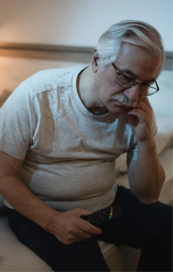 Older man seated with a television remote and a hand against his cheek.